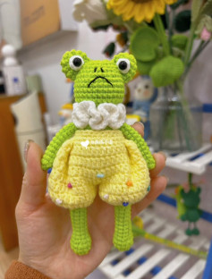 Crochet pattern of green frog with bulging eyes, wearing yellow pants.